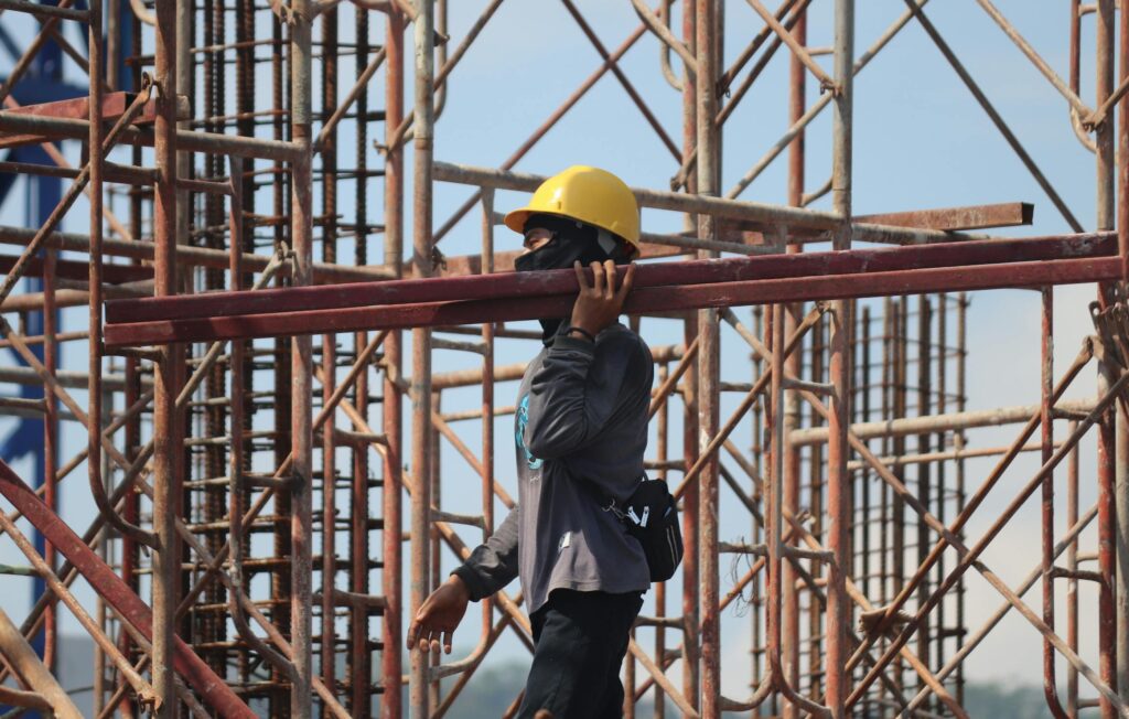 A construction worker with safety gear carrying a metal beam amidst scaffolding at a busy construction site.