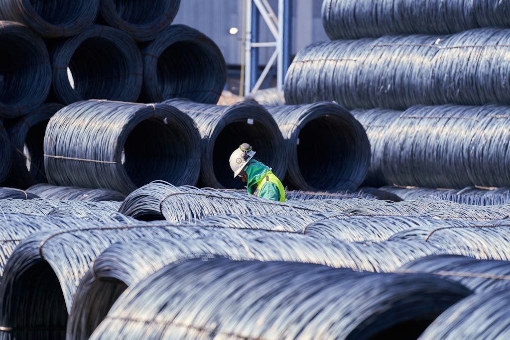 Construction worker in safety gear navigates large stacks of metal wire coils at an industrial site.