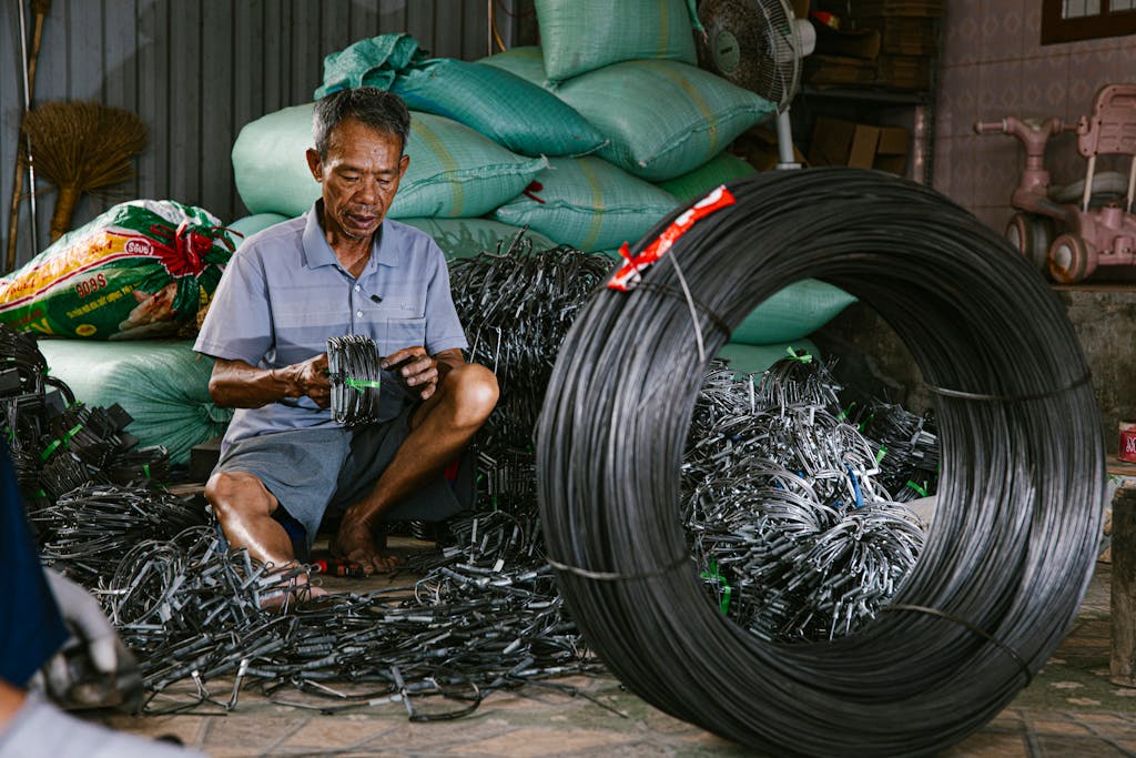 Elderly man seated among wire coils, methodically organizing materials in a workshop setting.