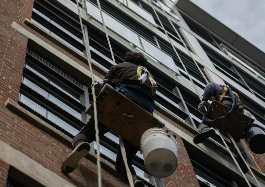 Two workers cleaning windows on a tall building using ropes. Urban setting, low angle shot.